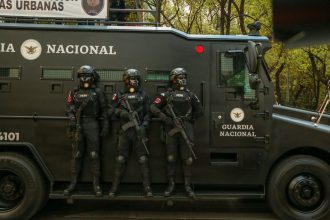 Agentes de segurança em equipamento tático ao lado de um veículo blindado durante uma operação.
