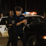 Policiais realizam uma operação noturna ao lado de uma viatura em uma rua pouco iluminada.
