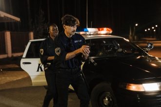 Policiais realizam uma operação noturna ao lado de uma viatura em uma rua pouco iluminada.