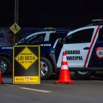 Barreira policial noturna com viaturas, cones e agentes realizando fiscalização em uma via urbana.