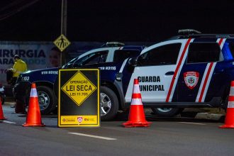Viaturas e agentes em um bloqueio policial noturno, com cones sinalizando a via.